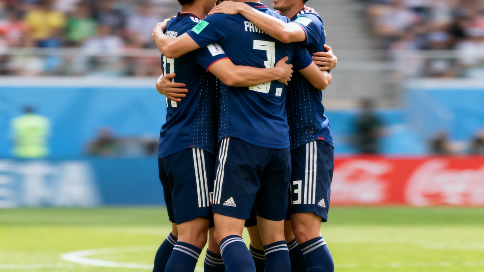 2026世界杯亚洲区预选赛日本队球员在进球后集体庆祝的抓拍照片，绿色草坪上四名球员拥抱在一起，背景是模糊的观众看台。